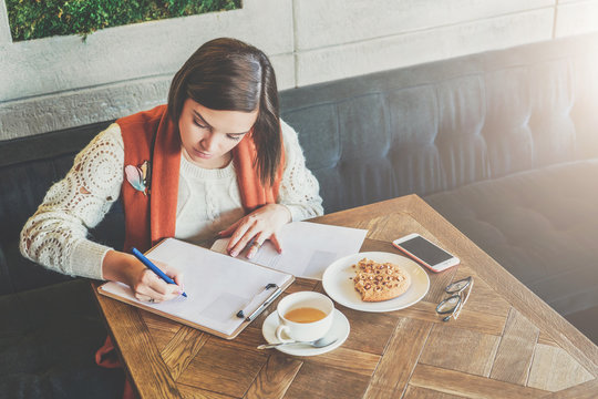 Young businesswoman is sitting in cafe at table, signing documents. Student is studying online, doing homework. Girl is writing letter, statement. On table cup of tea, cookies, smartphone. E-learning.