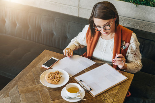 Young businesswoman in glasses and white sweater is sitting in cafe at table,signing documents,working. Student is studying online,doing homework. On table cup of tea, cookies, smartphone. E-learning.