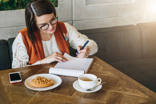 Young businesswoman in glasses and white sweater is sitting in cafe at table, working. Girl analyzes data, plans, considers income. Student learning online. On table cup of tea,smartphone. E-learning.