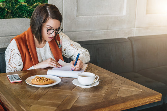 Young businesswoman in glasses and white sweater is sitting in cafe at table, working. Girl fills out an application, questionnaire, signs documents, draws up resume. Student is studying.E-learning.