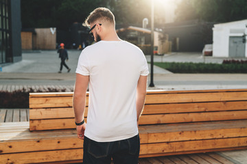 Summer day. Back view. Young bearded hipster man dressed in white t-shirt and sunglasses is stands...