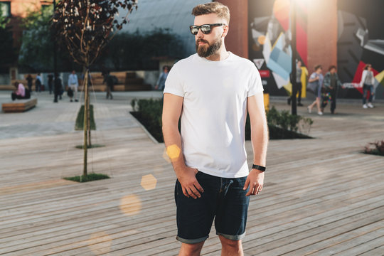 Summer day. Front view. Young bearded hipster man dressed in white t-shirt and sunglasses is stands on city street. Mock up. Space for logo, text, image. Instagram filter, film effect, bokeh effect.