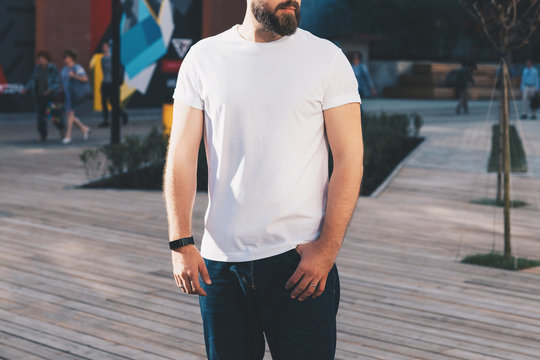 Summer day. Front view. Young bearded hipster man dressed in white t-shirt and sunglasses is stands on city street. Mock up. Space for logo, text, image. Instagram filter, film effect, bokeh effect.