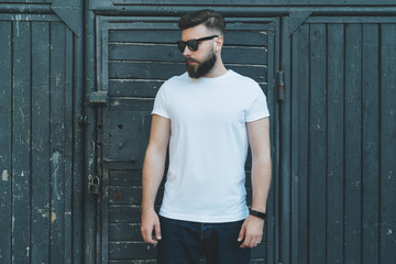 Summer day. Front view. Young bearded hipster man dressed in white t-shirt and sunglasses is stands...