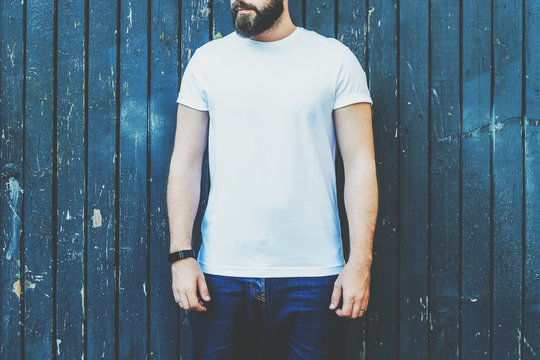 Summer day. Front view. Young bearded hipster man dressed in white t-shirt and sunglasses is stands against dark wood wall. Mock up. Space for logo, text, image.