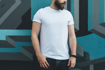 Summer day. Front view. Young bearded hipster man dressed in white t-shirt is stands against wall with graffiti. Mock up. Space for logo, text, image.