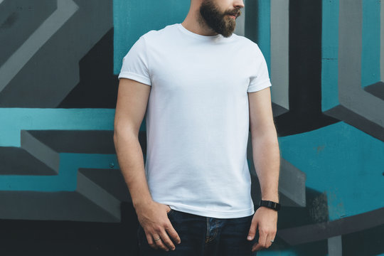 Summer day. Front view. Young bearded hipster man dressed in white t-shirt is stands against wall with graffiti. Mock up. Space for logo, text, image.