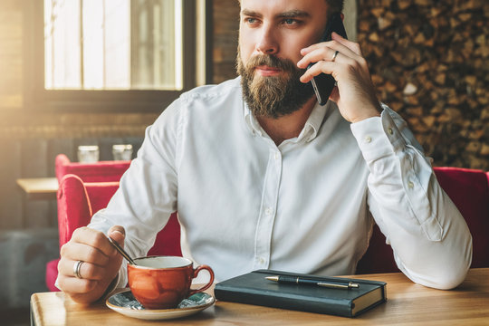 Young bearded businessman sits in cafe at table, drinking tea, talking on cellphone. On table notebook, pen. Man is working, studying.Online education, marketing, training.E-learning,e-commerce.