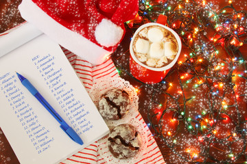 Santa's preparation for christmas presents delivery: winter hot spicy drink cacao with marshmallows and list of names naughty and nice children on background of striped blanket and electric lights