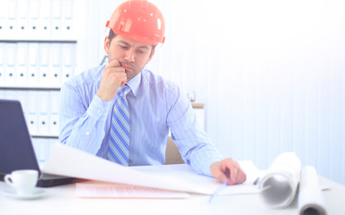 Architect looking working in office at desk. Wearing hardhat and taking notes on paper