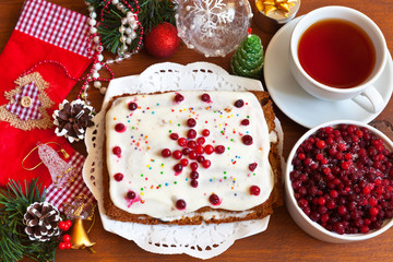 New Year's tea: homemade two-layered pie covered with milk cream and red bilberry in the form of snowflakes; a cup of black tea and a bowl of frozen berries on a wooden table. Christmas still life