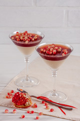 Creamy vanilla panna cotta with red jelly in beautiful glasses, fresh ripe pomegranate on white wooden background.