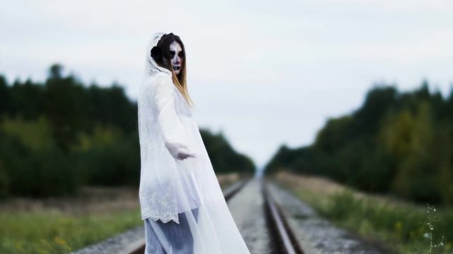 The young woman with make-up of dead bride for Halloween dressed in white wedding gown is going on the railway track among the autumn forest. 4K