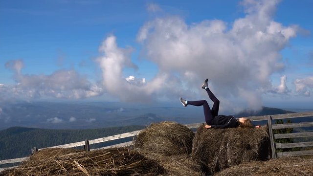 a young girl resting in the Alpine mountains lying on the haystack turns the legs on a background of blue sky with clouds. in the background panorama of mountains and wood fence