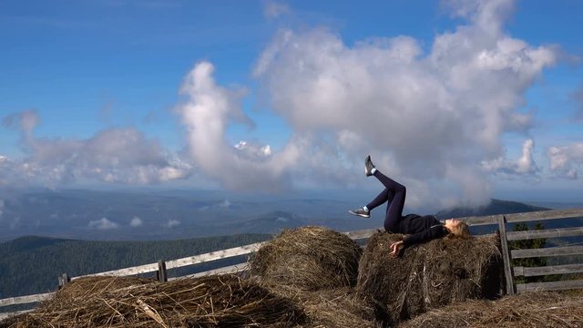 a young girl resting in the Alpine mountains lying on the haystack turns the legs on a background of blue sky with clouds. in the background panorama of mountains and wood fence