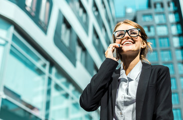 Young blonde caucasian businesswoman in a white blouse and a black jacket is talking phone before the modern corporation building in the background