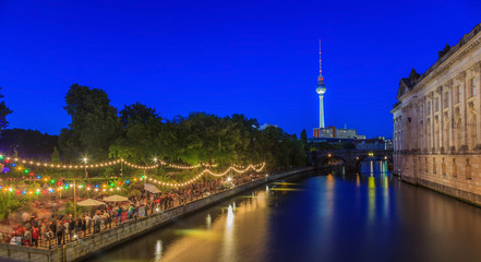 Aufnahme eines Biergartens am Berliner Spreeufer gegenüber des Bode Museums Abends zur Dämmerung in der blauen Stunde mit Fernsehturm im Hintergrund und Lichtspiegelungen in der Spree © Aquarius