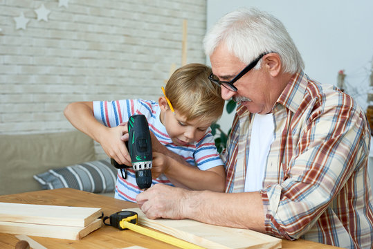 Portrait of grandfather helping little boy build birdhouse, teaching him woodwork at table in workshop