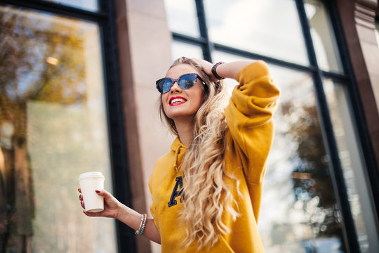 young beautiful blonde drinking coffee walking around the city.mustard sweetshot.,urban backpack , bright red lips Posing against the window of the boutique
