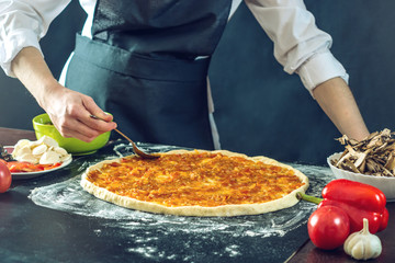 The chef in black apron makes pizza with his hands putting the ingredients for the pizza on the table.