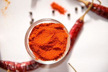 A glass bowl filled with bright red chili powder, surrounded by dried red chilies and black pepper on a white background. The fine ground spice adds a bold, fiery flavor to various culinary dishes