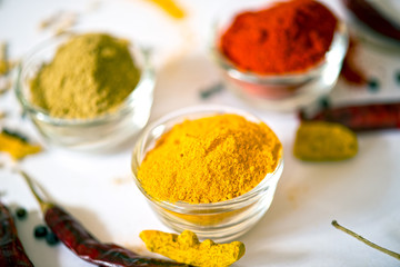 A close-up of three glass bowls filled with chili powder, turmeric powder, and coriander powder, surrounded by dried red chilies, black peppercorns, and turmeric roots on a white surface