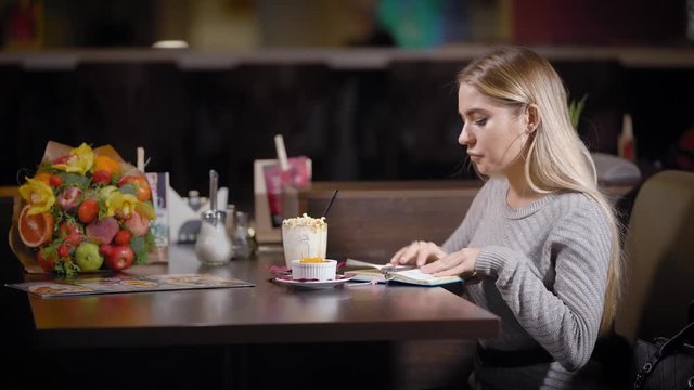 A young and pretty woman is eating lunch in a restaurant, a lady eating a dessert with milkshakes, before that she wrote down her thoughts