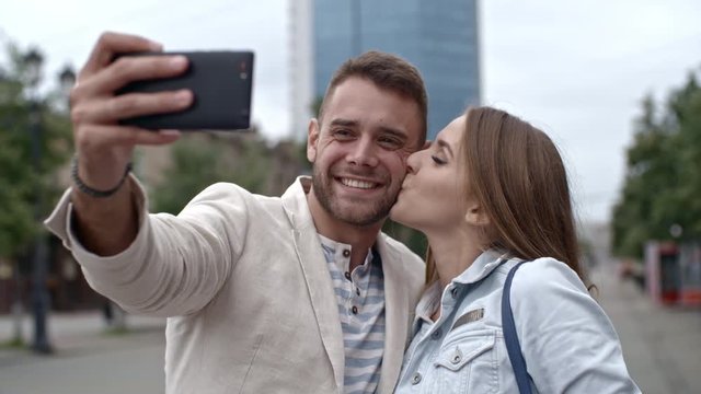 Loving young couple embracing and kissing at camera while taking selfie with smartphone on the street