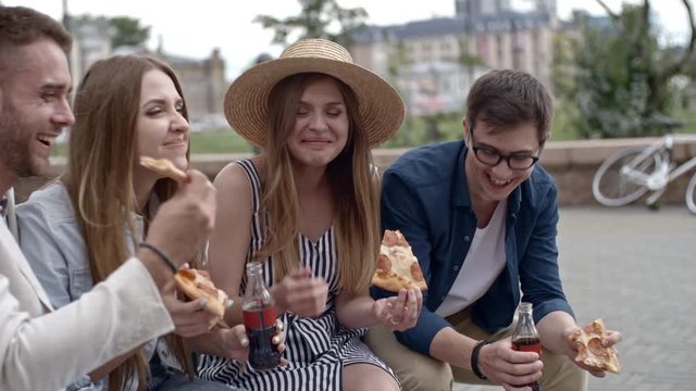 Young couples drinking coke from glass bottles, eating pizza, talking and laughing while sitting on bench outdoors