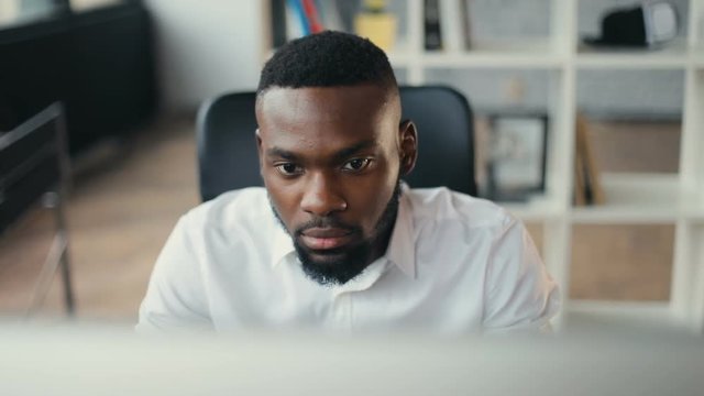African American handsome male uses computer for work in modern office programmer serious looks at monitor screen freelance successful worker businessman solves engineering problems Slow motion