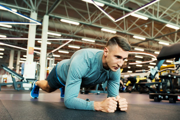 Fototapeta na wymiar Portrait of fit young man doing plank excursive on floor in modern gym, looking away with determination
