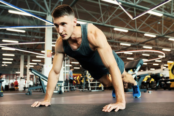 Naklejka na ściany i meble Portrait of handsome young man with muscular arms doing push ups on floor in modern gym