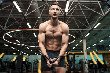 Fototapeta na wymiar Low angle portrait of determined young man with bare chest pumping muscles working out on machines in gym in dramatic light