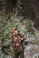 Fototapeta premium Male hunter with backpack, armed with a rifle, standing in a snowy winter forest. Man looks away. Snowfall in the forest.