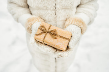 Female hand in a white knitted mittens holding craft paper gift box with as a present for Christmas, new year, valentine day or anniversary on winter background.