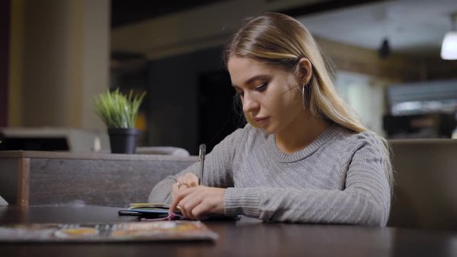A cute and young woman who sits in a cafe writes poetry in a notebook, a lady holds a pen in her hand for notes of her thoughts and composing new lines