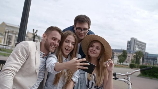 Group of young joyful women and men sitting together on bench on city street, smiling and posing at camera of smartphone while taking a selfie