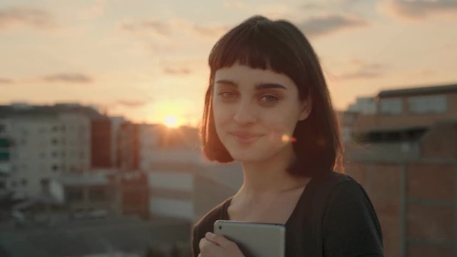 Playful beautiful young woman in her 20s, with short haircut and trendy hipster tattoos stands in warm summer sunset light, holds tablet device, chuckles and smiles into camera, shows tongue