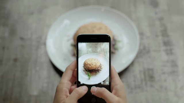 Top view of food blogger or social media influencer makes photos of her amazing huge mouth watering fast food burger on smartphone, obsessed with publishing online