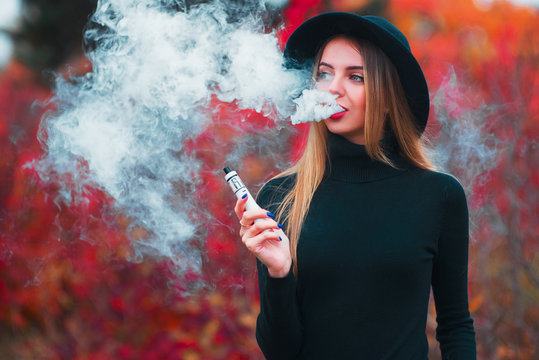 Vaping. Young beautiful woman in hat vaping outdoors in autumn