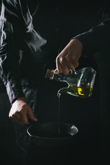 Man chef in black apron pouring olive oil from bottle for cooking pancakes in cast-iron pan. Dark rustic style. Toned image