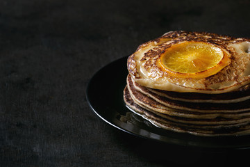 Stack of homemade pancakes with fried orange, served with mapple syrup on black plate over dark background. Close up