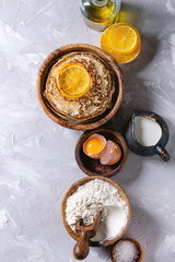 Homemade pancakes with fried orange and ingredients above. Wooden bowls of flour, yolk, salt, milk, olive oil over gray texture background. Top view with space. Home cooking concept