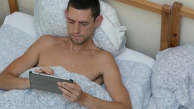 Young man is typing something on a lepton or tablet lying on the bed stretching and yawning