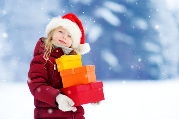 Adorable little girl wearing Santa hat holding a pile of Christmas gifts on beautiful winter day