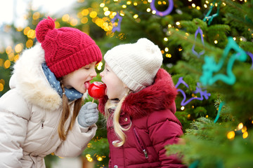 Two adorable little sisters eating red apples covered with sugar icing on traditional Christmas...