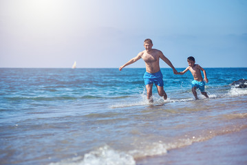 Caucasian dad and son are running in the water along the sea shore.
