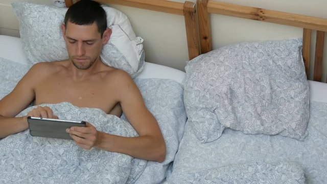Young man is typing something on a lepton or tablet lying on the bed stretching and yawning