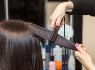 The hairdresser does aligns the hair with hair iron to a young girl, brunette in a beauty salon. Professional Hair Care