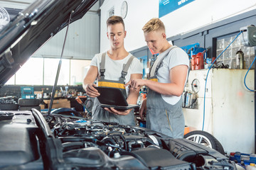 Experienced auto mechanic using a laptop for scanning and interpreting engine error codes next to an apprentice in a modern automobile repair shop